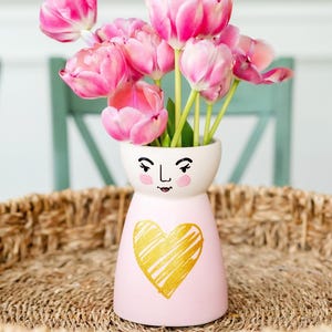May include: Pink ceramic vase with a hand-painted face and a gold heart design. The vase is filled with pink tulips.