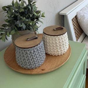 May include: Two crocheted storage baskets with wooden lids and leather handles. The baskets are grey and white and are sitting on a wooden tray on a green dresser.