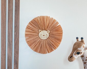 May include: A plush giraffe toy with brown and white spots stands next to a blue velvet cushion. A brown and beige woven lion head wall hanging is on the wall behind the cushion.