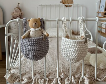 Op de afbeelding: Twee witte metalen hangmanden met grijze en witte gehaakte stoffen voeringen. De manden hangen aan een wit metalen bedframe met wit en roze beddengoed met bloemenprint.
