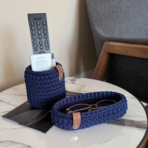 Puede incluir: Dos cestas de crochet azules, una con un mando a distancia y un bolígrafo, la otra con unas gafas. Las cestas tienen una etiqueta de cuero marrón con la palabra "Amitié" en ellas. Las cestas están sobre una mesa de mármol blanco con un borde dorado.