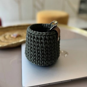 Può includere: Un portapenne in crochet verde scuro con un cinturino in pelle marrone. Il portapenne è appoggiato su un laptop argentato.