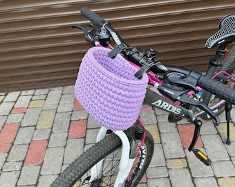 Puede incluir: Una cesta de bicicleta de ganchillo lavanda sujeta al manillar de una bicicleta de montaña negra y rosa. La cesta es cilíndrica y tiene un aspecto texturizado. La bicicleta tiene manillar negro y horquilla delantera blanca. El fondo es una puerta de garaje marrón.