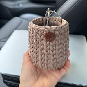Pode incluir: Um recipiente de forma cilíndrica de cor bege, feito de crochê, com uma etiqueta de couro marrom. O recipiente contém um par de óculos.