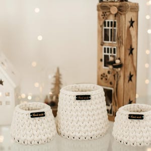 May include: Three white crocheted baskets with black "Hand Made" labels on a white surface. The baskets are of different sizes and are arranged in a row.