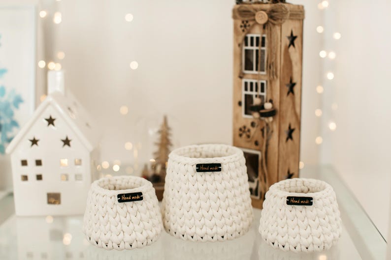 May include: Three white crocheted baskets with black "Hand Made" labels on a white surface. The baskets are of different sizes and are arranged in a row.