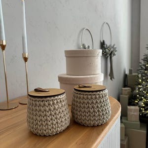 Puede incluir: Dos contenedores de almacenamiento de crochet beige con tapas de madera. Los contenedores est&aacute;n sobre una superficie de madera.