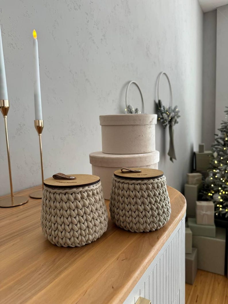 Puede incluir: Dos contenedores de almacenamiento de crochet beige con tapas de madera. Los contenedores est&aacute;n sobre una superficie de madera.