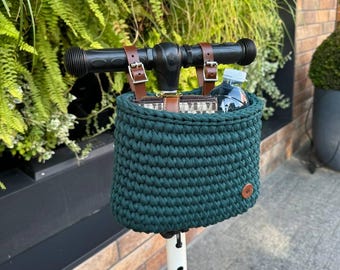 May include: A green crocheted basket with brown leather straps is attached to the handlebars of a white scooter. A water bottle is inside the basket.