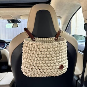 May include: A beige crocheted hanging basket with brown leather straps. The basket is hanging from the headrest of a car seat.