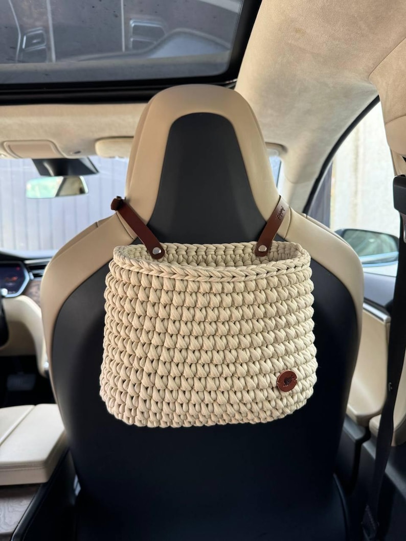 May include: A beige crocheted hanging basket with brown leather straps. The basket is hanging from the headrest of a car seat.