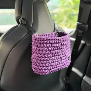 May include: A handcrafted, lavender-colored crochet car organizer. The basket-shaped organizer hangs from the car seat headrest with a black strap. The interior is visible, and the texture of the crochet is prominent.