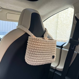 Pu&ograve; includere: Borsa organizer per auto all'uncinetto beige appesa a un poggiatesta per auto. La borsa &egrave; rettangolare con una trama intrecciata e una piccola etichetta nera. Il cinturino &egrave; realizzato con lo stesso materiale all'uncinetto beige.
