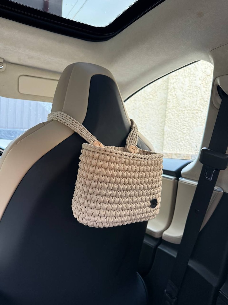 Pu&ograve; includere: Borsa organizer per auto all'uncinetto beige appesa a un poggiatesta per auto. La borsa &egrave; rettangolare con una trama intrecciata e una piccola etichetta nera. Il cinturino &egrave; realizzato con lo stesso materiale all'uncinetto beige.