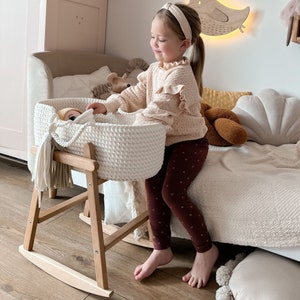 Puede incluir: Un moisés blanco tejido con un soporte de madera para mecer. Una niña está sentada en una cama y sostiene una muñeca dentro del moisés.