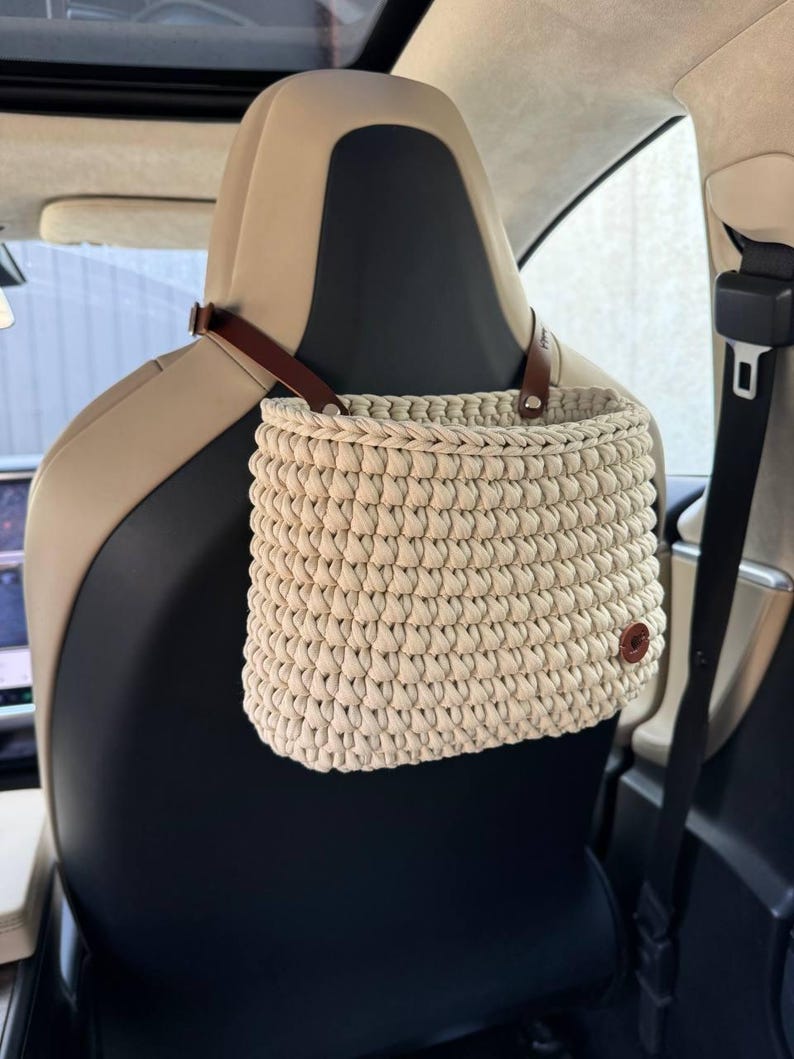 May include: A beige crocheted hanging basket with brown leather straps, hanging from the headrest of a car seat.