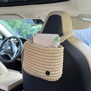 Pu&ograve; includere: Organizzatore per auto all'uncinetto beige appeso al poggiatesta del sedile dell'auto. L'organizzatore ha una trama intrecciata e una piccola etichetta nera rotonda. All'interno sono visibili un tablet e una bottiglia d'acqua. L'interno dell'auto &egrave; visibile.