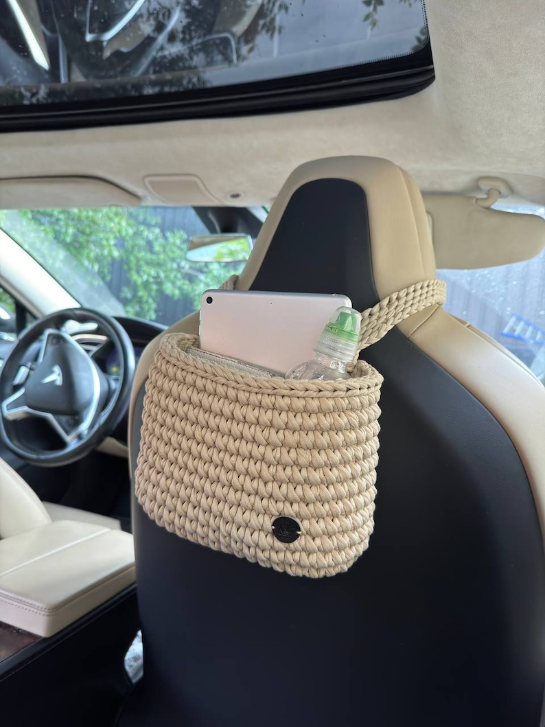Pu&ograve; includere: Organizzatore per auto all'uncinetto beige appeso al poggiatesta del sedile dell'auto. L'organizzatore ha una trama intrecciata e una piccola etichetta nera rotonda. All'interno sono visibili un tablet e una bottiglia d'acqua. L'interno dell'auto &egrave; visibile.