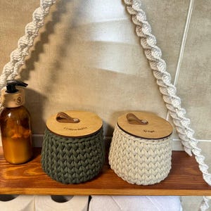Puede incluir: Dos cestas de almacenamiento de crochet con tapas de madera y asas de cuero. La cesta verde está etiquetada como "Cotton Balls" y la cesta beige está etiquetada como "Q-tips".