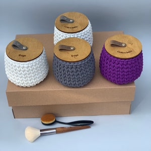 Puede incluir: Tres contenedores de crochet con tapas de madera, etiquetados como "Cotton Balls", "Q-tips" y "Q-tips". Los contenedores son blancos, grises y morados. Dos pinceles de maquillaje est&aacute;n sobre una caja marr&oacute;n debajo de los contenedores.