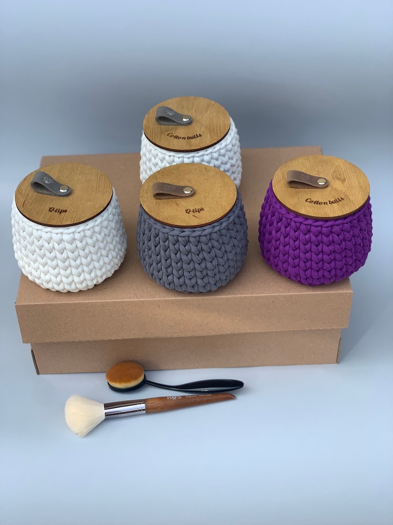 Puede incluir: Tres contenedores de crochet con tapas de madera, etiquetados como "Cotton Balls", "Q-tips" y "Q-tips". Los contenedores son blancos, grises y morados. Dos pinceles de maquillaje est&aacute;n sobre una caja marr&oacute;n debajo de los contenedores.