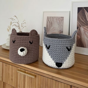 May include: Two crocheted storage baskets with animal faces. The brown basket has a bear face and the gray basket has a wolf face. The baskets are sitting on a wooden surface.