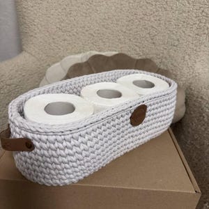 Könnte beinhalten: Ein hellgrauer, ovaler Häkelkorb mit drei Rollen weißem Toilettenpapier. Der Korb hat braune Lederakzente und -griffe. Er steht auf einer braunen Pappschachtel.