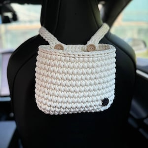 May include: White crocheted hanging basket with two buttons on the straps. The basket is hanging from the headrest of a car seat.