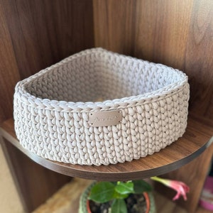 May include: A beige crocheted storage basket with a small metal tag that says "Aminika" on a wooden shelf.