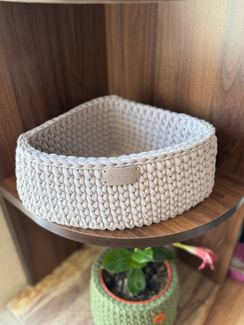 May include: A beige crocheted storage basket with a small metal tag that says "Aminika" on a wooden shelf.