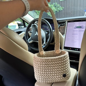 Pu&ograve; includere: Borsa tote beige lavorata all'uncinetto a mano con due manici intrecciati e dettagli con bottoni in legno. La borsa &egrave; cilindrica con una trama intrecciata e una piccola etichetta circolare nera. La borsa &egrave; mostrata all'interno di un'auto.