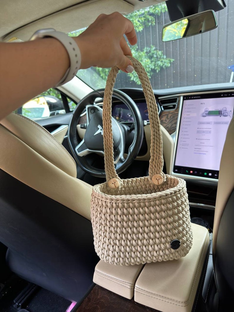Pu&ograve; includere: Borsa tote beige lavorata all'uncinetto a mano con due manici intrecciati e dettagli con bottoni in legno. La borsa &egrave; cilindrica con una trama intrecciata e una piccola etichetta circolare nera. La borsa &egrave; mostrata all'interno di un'auto.