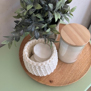 Puede incluir: Una cesta de crochet blanca llena de discos de algod&oacute;n se encuentra sobre una bandeja de madera con un frasco de vidrio con tapa de madera. Una planta verde en maceta est&aacute; en el fondo.