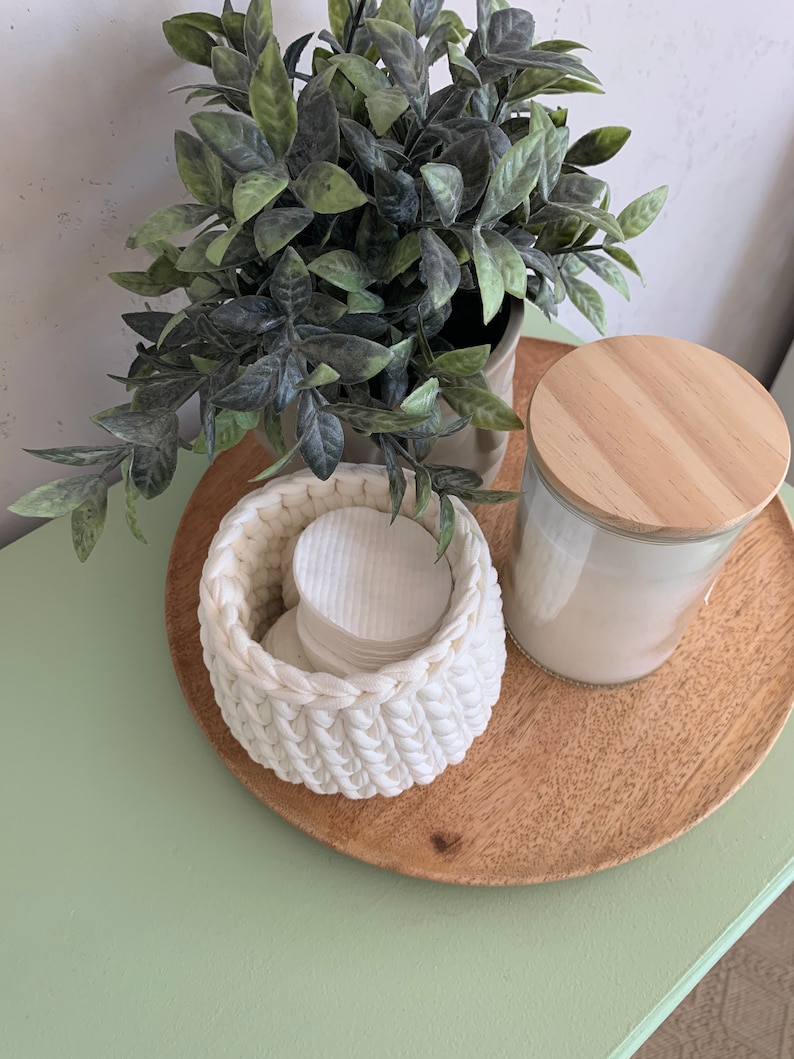 Puede incluir: Una cesta de crochet blanca llena de discos de algod&oacute;n se encuentra sobre una bandeja de madera con un frasco de vidrio con tapa de madera. Una planta verde en maceta est&aacute; en el fondo.