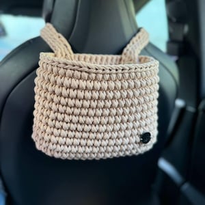 May include: A beige crocheted hanging storage basket with a black button. The basket is hanging from the headrest of a car seat.