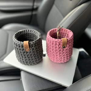 Peut inclure: Deux paniers de rangement en crochet, l'un gris et l'autre rose, avec des étiquettes en cuir qui disent "GIBD". Les paniers sont posés sur une surface grise.
