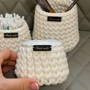 May include: Three white crocheted baskets with black leather tags that say "Hand made". The smallest basket is filled with cotton buds, the middle basket is filled with makeup brushes, and the largest basket is empty.