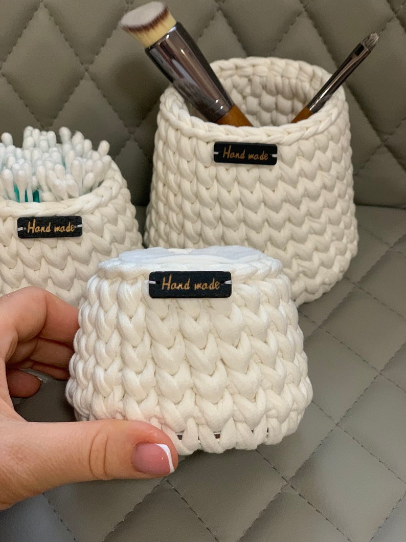 May include: Three white crocheted baskets with black leather tags that say "Hand made". The smallest basket is filled with cotton buds, the middle basket is filled with makeup brushes, and the largest basket is empty.