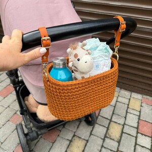 Op de afbeelding: Een bruine gehaakte mand met een leren riem bevestigd aan een kinderwagen. De mand bevat een blauwe waterfles, een knuffelgiraf en een luier.