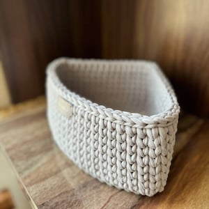 May include: A beige crocheted basket with a woven texture. The basket is empty and sitting on a wooden surface.