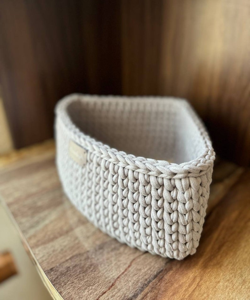May include: A beige crocheted basket with a woven texture. The basket is empty and sitting on a wooden surface.