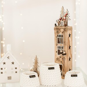 May include: Three white crocheted baskets with wooden tags that say "Hand Made" on a glass shelf. The baskets are of different sizes and are arranged in a row. A wooden Christmas house with a "Snow" sign is in the background.