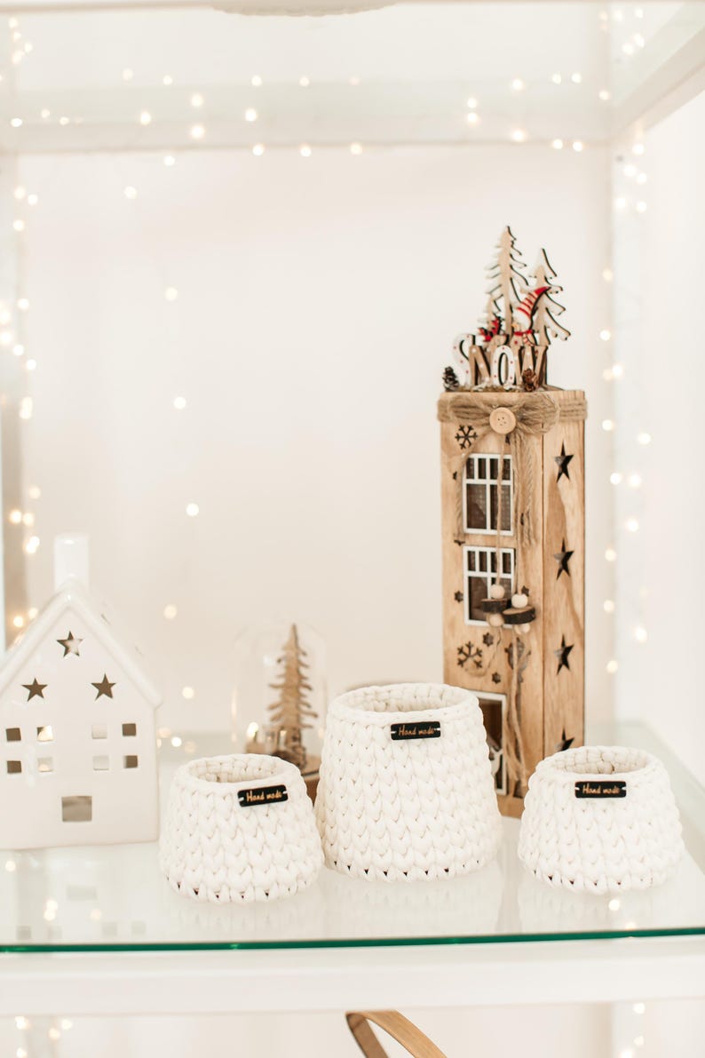 May include: Three white crocheted baskets with wooden tags that say "Hand Made" on a glass shelf. The baskets are of different sizes and are arranged in a row. A wooden Christmas house with a "Snow" sign is in the background.