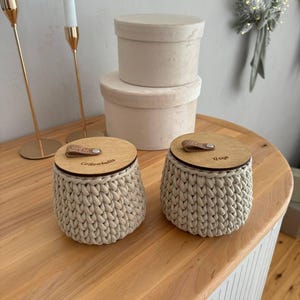Puede incluir: Dos contenedores de almacenamiento de crochet beige con tapas de madera. Las tapas tienen asas de cuero y est&aacute;n grabadas con las palabras "Glitterballs" y "Quip".