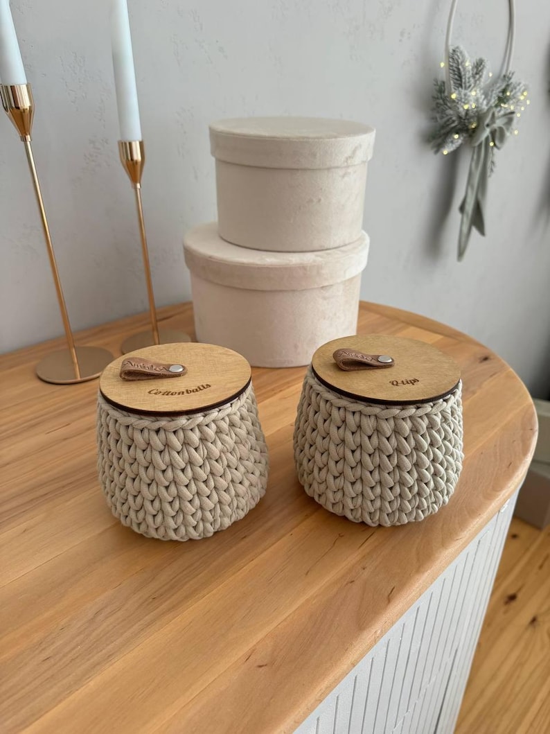 Puede incluir: Dos contenedores de almacenamiento de crochet beige con tapas de madera. Las tapas tienen asas de cuero y est&aacute;n grabadas con las palabras "Glitterballs" y "Quip".