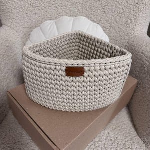 May include: A light beige, triangular crochet basket with a brown leather tag that reads "Annika". The basket is sitting on a brown cardboard box. The basket is made of thick yarn and has a braided edge.