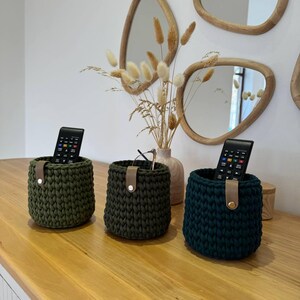 Puede incluir: Tres cestas de crochet en verde y verde oscuro con asas de cuero y botones metálicos. Las cestas están llenas de mandos a distancia y gafas. Las cestas están sobre una superficie de madera.