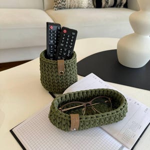 Puede incluir: Dos mandos a distancia negros están en una cesta de crochet verde. Una cesta ovalada de crochet verde con una etiqueta de cuero marrón está sobre una superficie blanca con un cuaderno y unas gafas dentro.