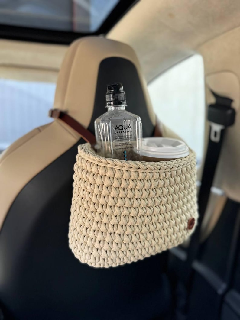 May include: A beige crocheted car seat organizer with a water bottle and a cup inside. The organizer is hanging from the headrest of a car seat.