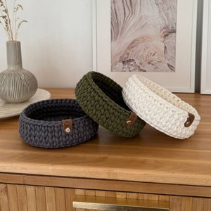 Può includere: Tre cesti in crochet di diversi colori: grigio, verde e bianco. I cesti sono realizzati con filato spesso e hanno una forma rotonda. Ogni cesto ha un bottone in pelle marrone sul lato.